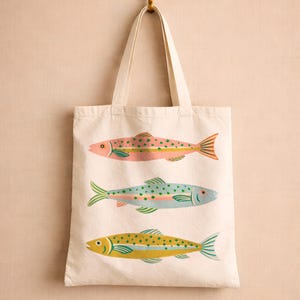May include: A cream-colored canvas tote bag featuring a screen-printed design of three fish stacked vertically. The top fish is pink with green spots, the middle fish is light blue with green spots, and the bottom fish is yellow with green spots.