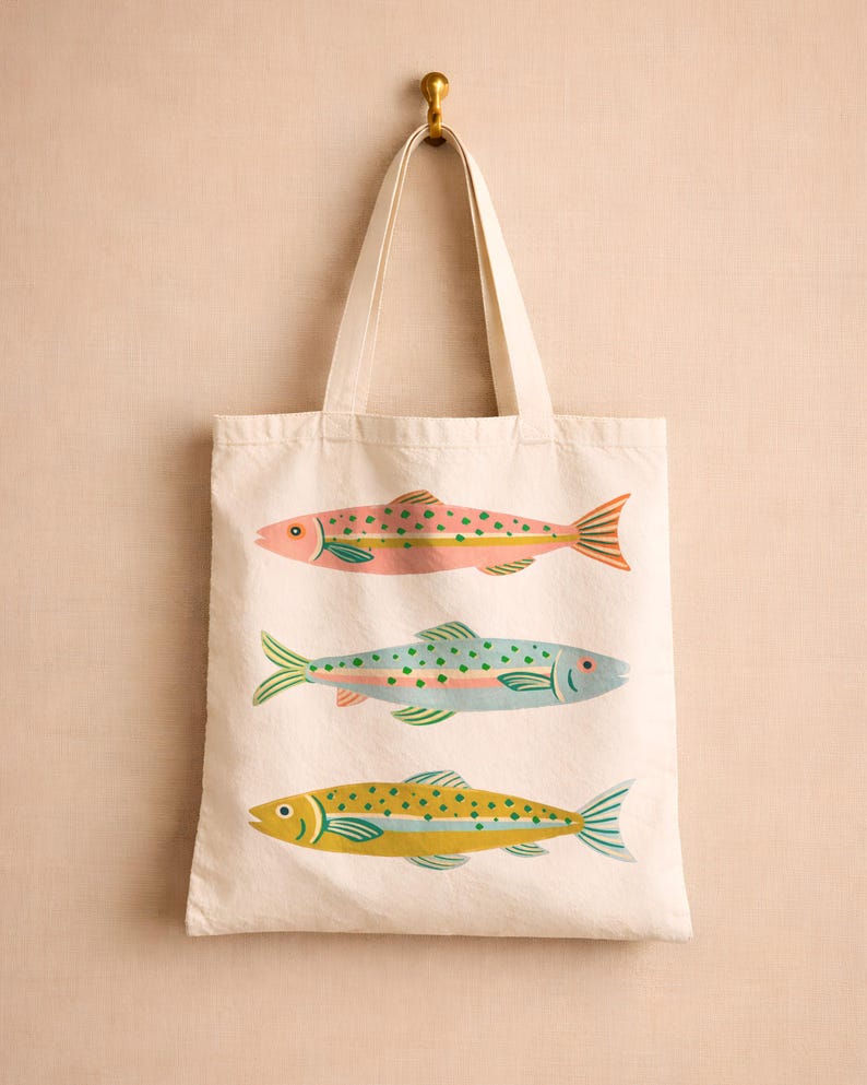 May include: A cream-colored canvas tote bag featuring a screen-printed design of three fish stacked vertically. The top fish is pink with green spots, the middle fish is light blue with green spots, and the bottom fish is yellow with green spots.