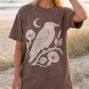 May include: A brown short-sleeve t-shirt featuring a large cream-colored graphic print of a raven perched on a flowering branch with a crescent moon above. The design is centered on the chest, showcasing detailed line work of the bird and flowers.