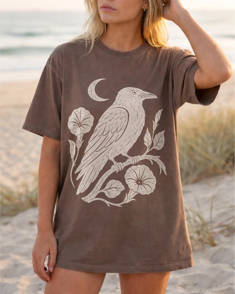 May include: A brown short-sleeve t-shirt featuring a large cream-colored graphic print of a raven perched on a flowering branch with a crescent moon above. The design is centered on the chest, showcasing detailed line work of the bird and flowers.