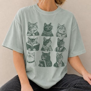 May include: A sage green short-sleeve t-shirt featuring a dark green graphic print of nine distinct cats arranged in a grid. Each cat is depicted in a different outfit or activity, including wearing scarves, sweaters, ties, and glasses, or reading books.