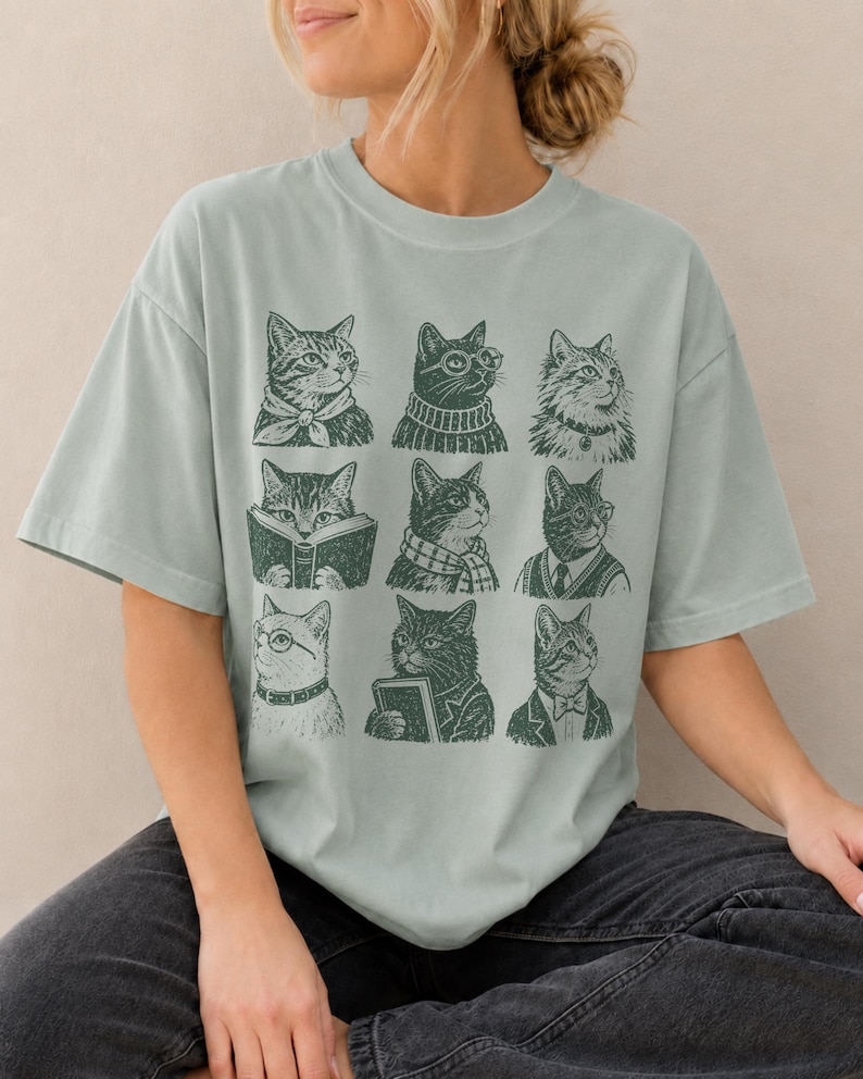 May include: A sage green short-sleeve t-shirt featuring a dark green graphic print of nine distinct cats arranged in a grid. Each cat is depicted in a different outfit or activity, including wearing scarves, sweaters, ties, and glasses, or reading books.