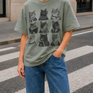 May include: A sage green short-sleeve t-shirt featuring a black graphic print of nine distinct cats arranged in a grid. Each cat is depicted with different accessories or clothing items, including glasses, scarves, sweaters, and books, in a detailed line art style.