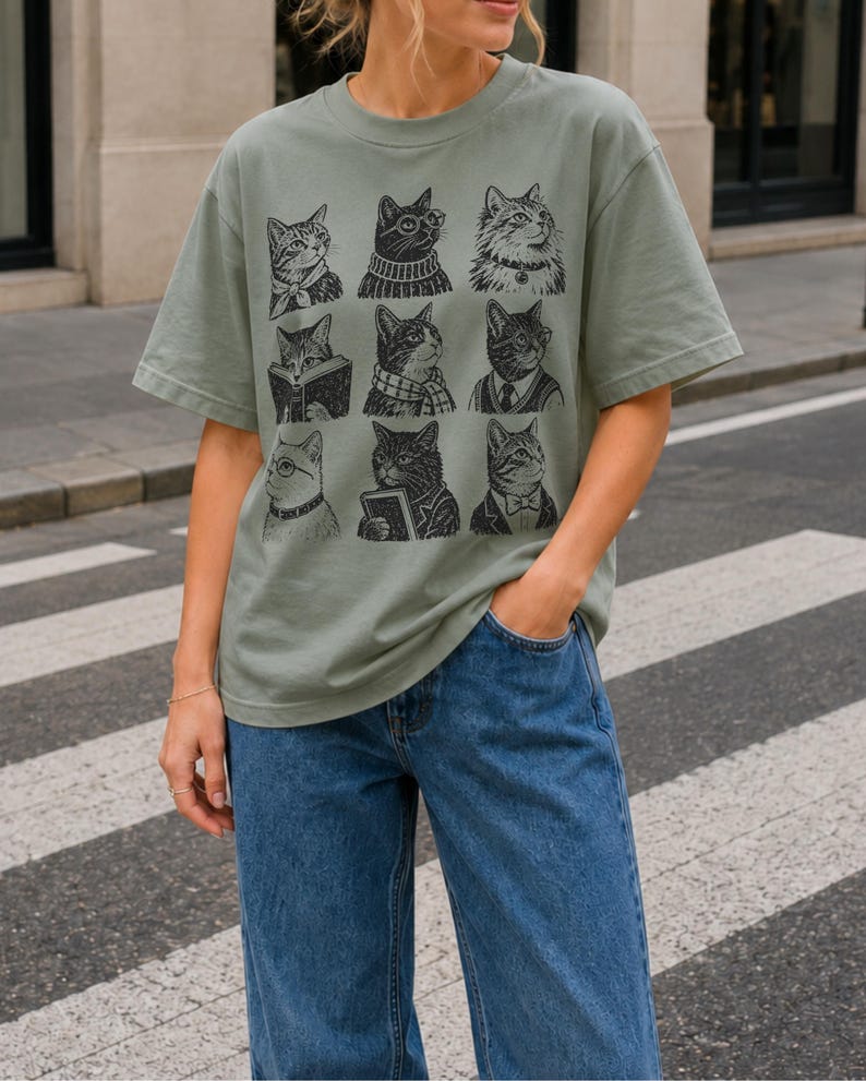 May include: A sage green short-sleeve t-shirt featuring a black graphic print of nine distinct cats arranged in a grid. Each cat is depicted with different accessories or clothing items, including glasses, scarves, sweaters, and books, in a detailed line art style.
