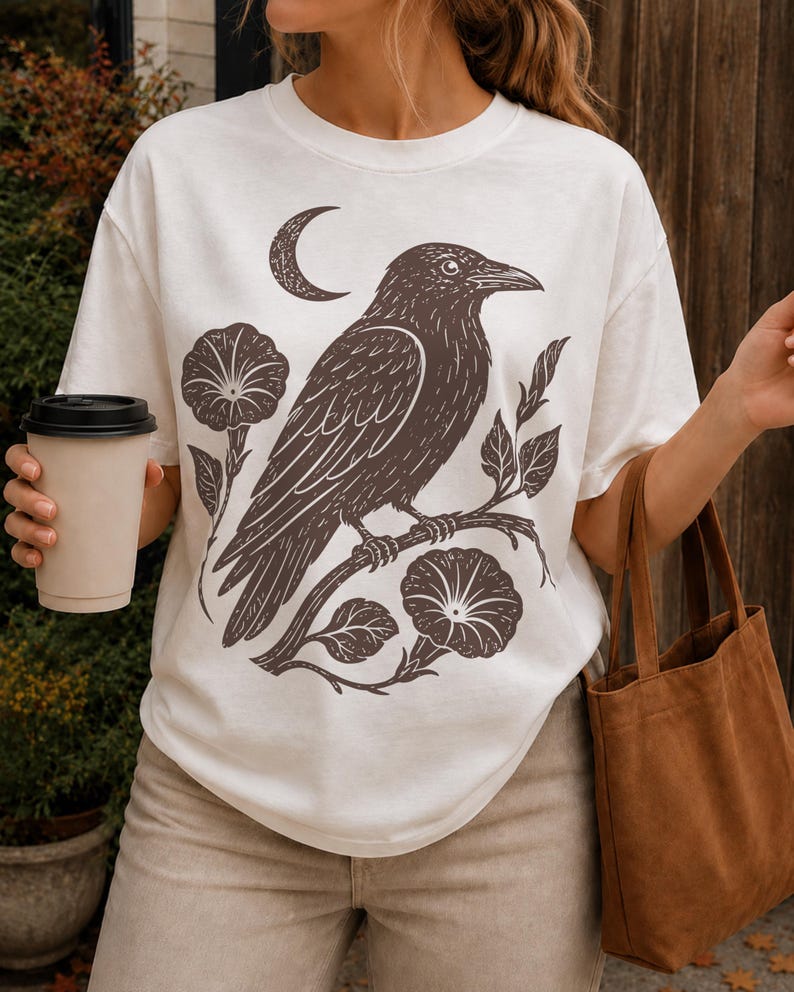 May include: A white short-sleeved t-shirt featuring a dark brown graphic print of a crow perched on a flowering branch with a crescent moon above. The design includes detailed line work of the bird, leaves, and two large open blossoms.