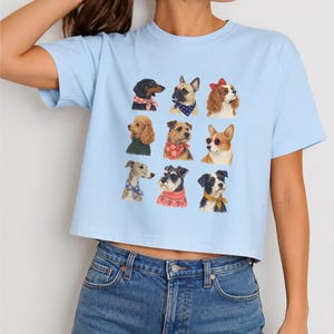 Puede incluir: Una camiseta de manga corta de color azul claro con un estampado gr&aacute;fico de nueve razas de perros diferentes dispuestas en cuadr&iacute;cula. Cada perro est&aacute; ilustrado con accesorios distintos, como pa&ntilde;uelos estampados, un lazo rojo y gafas de sol.
