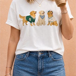 May include: A white short-sleeved t-shirt featuring a graphic print of three golden retriever dogs. The first dog wears a green sweater with a letter G, the second wears a blue floral bandana, and the third wears a white sweater and green cap.