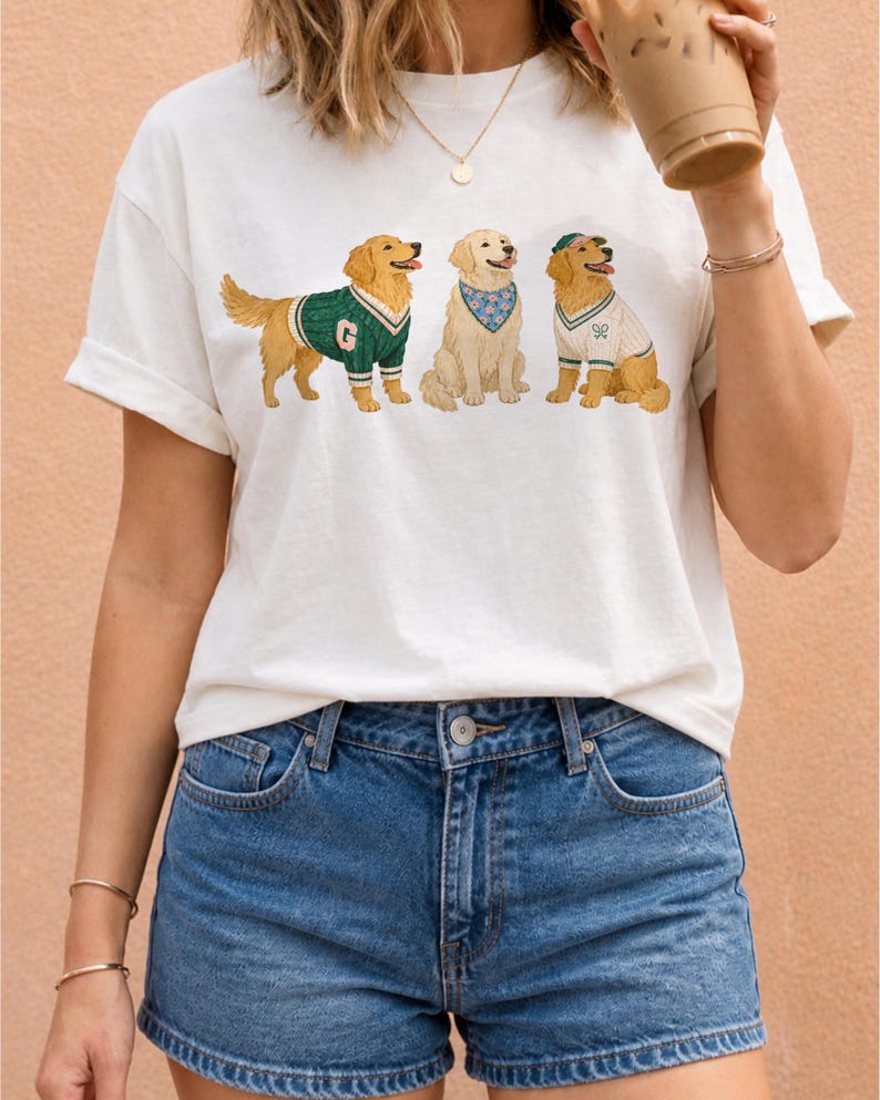 May include: A white short-sleeved t-shirt featuring a graphic print of three golden retriever dogs. The first dog wears a green sweater with a letter G, the second wears a blue floral bandana, and the third wears a white sweater and green cap.
