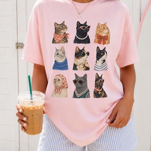 以下が含まれることがあります： 淡いピンク色の半袖Tシャツで、9匹の猫のイラストがグリッド状に配置されています。それぞれの猫は、スカーフ、サングラス、パールネックレス、リボンなどの異なるアクセサリーを身につけており、前面中央に3列ずつ並んでいます。