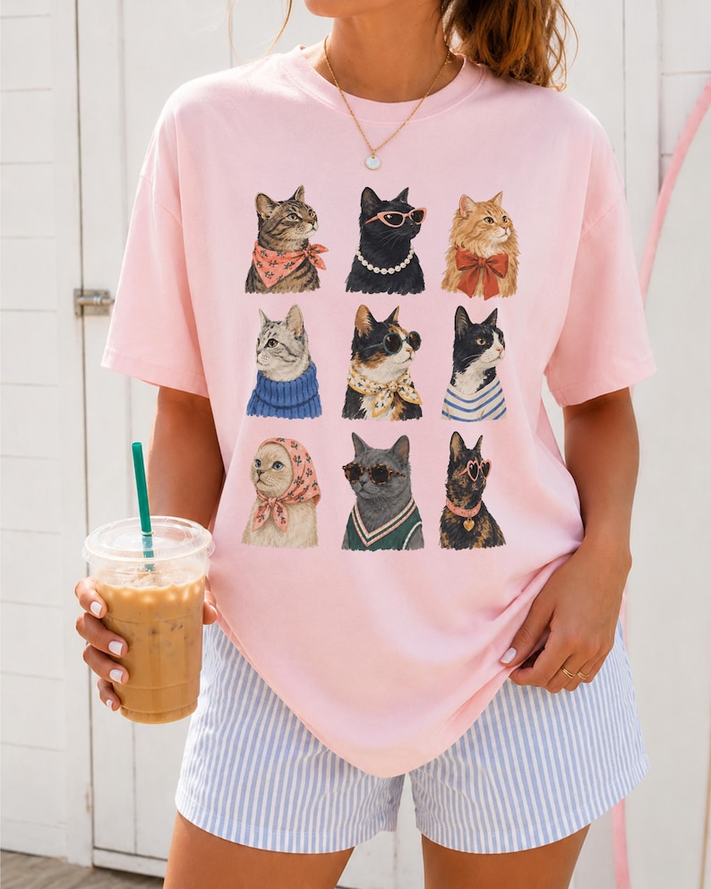 以下が含まれることがあります： 淡いピンク色の半袖Tシャツで、9匹の猫のイラストがグリッド状に配置されています。それぞれの猫は、スカーフ、サングラス、パールネックレス、リボンなどの異なるアクセサリーを身につけており、前面中央に3列ずつ並んでいます。