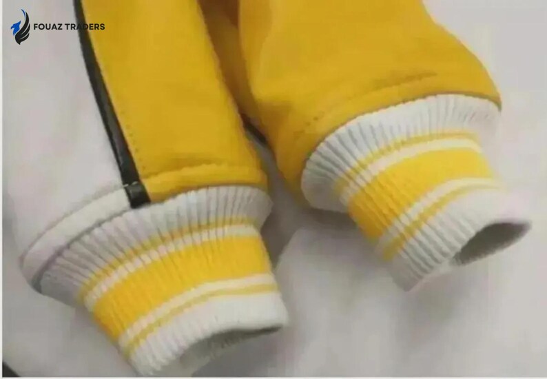 Puede incluir: Primer plano de una manga de chaqueta amarilla con pu&ntilde;os acanalados a rayas blancas y amarillas. La chaqueta tiene un panel blanco con una raya negra. La marca "FOUAZ TRADERS" es visible.