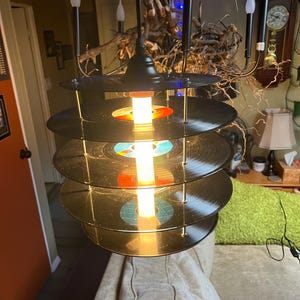 May include: A black chandelier made from stacked vinyl records with a single light bulb in the center. The chandelier has five candle-style light bulbs.