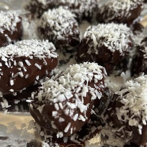 May include: Close-up of chocolate-covered treats sprinkled with shredded coconut. The treats are arranged on a silver surface. The chocolate coating is dark brown, and the coconut flakes are white.
