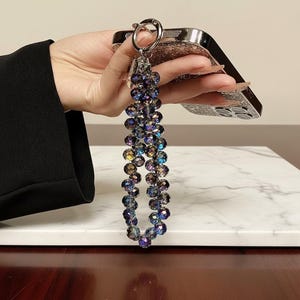 Könnte beinhalten: Ein Perlen-Telefonanhänger mit schillernden, mehrfarbigen Perlen. Der Anhänger ist an einem silberfarbenen Ring und einer Handyhülle mit glitzerndem Design befestigt. Die Perlen sind vertikal angeordnet.