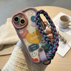 May include: A clear phone case with a lilac border, decorated with heart and smiley face stickers. The case has a braided strap in teal and purple. The words "Smile" and "Happy Day" are visible on the case.