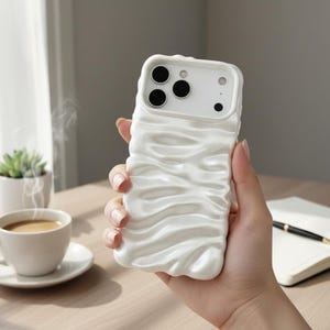 May include: A white phone case with a textured, wave-like pattern. The case has a glossy finish and cutouts for the phone's camera and buttons. The case is held in a hand, with a cup of coffee and a notebook visible in the background.