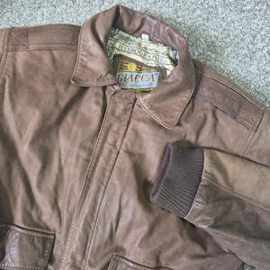May include: A brown leather jacket featuring a classic collar, ribbed knit cuffs, and two front pockets. The interior label displays the text GIACCA traditional and AC HYDROGRAPHICA. The jacket has reinforced shoulder panels and a button-front closure.