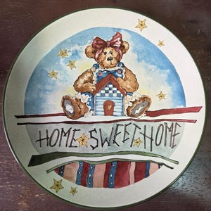Puede incluir: Un plato decorativo con una ilustración en acuarela. El diseño incluye un oso de peluche con un lazo y un peto, sosteniendo una casa. Las palabras "HOME SWEET HOME" están escritas en el plato, con acentos de estrellas. El plato tiene un borde verde.