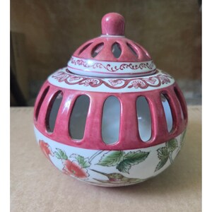 May include: A decorative ceramic jar with a lid. The jar is primarily white with a pink band around the middle and a pink lid. The jar has a floral design with red flowers and green leaves. The lid has a knob on top and decorative cutouts.