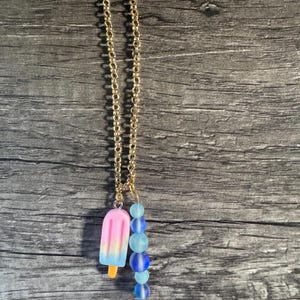 May include: A gold-colored chain necklace with a popsicle charm and a cluster of blue beads. The popsicle charm is pink, blue, and yellow. The beads are various shades of blue.