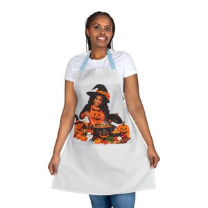May include: A white apron featuring a graphic print of a person wearing a witch hat and orange apron, stirring a black pot decorated with a jack-o'-lantern face. The scene includes pumpkins, a small ghost, and autumn leaves on a light background.