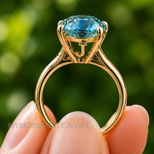 Puede incluir: Un anillo de oro con una gran piedra preciosa azul redonda y facetada en un engaste de cesta de perfil alto. El lateral de la galería de oro muestra una letra J prominente. El aro es liso y pulido, con una construcción metálica clásica.
