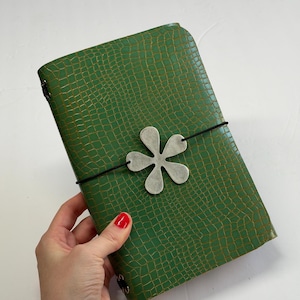 May include: A green journal with a textured crocodile skin pattern. The cover is secured by a black elastic band featuring a silver-toned, six-petaled flower charm centered on the front. The notebook has rounded corners and a slim, rectangular shape.