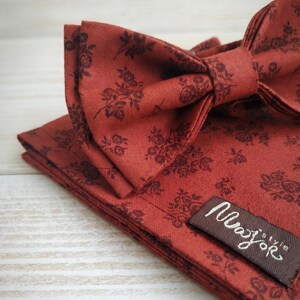May include: A rust-colored bow tie and pocket square set with a floral pattern. The bow tie is tied and sits atop the folded pocket square. A small brown tag with the text "Major style" is visible.