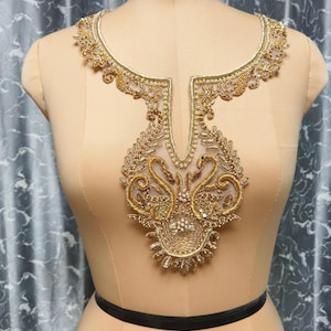 Pode incluir: Uma aplicação de decote bordada em dourado com dois motivos de cisnes, missangas intrincadas e detalhes em fio metálico sobre uma base de malha transparente. O design inclui uma abertura central e padrões florais ornamentais.