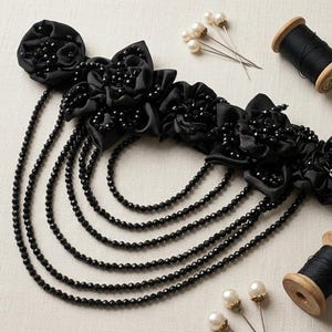 Può includere: Un'applicazione floreale in tessuto nero composta da diversi fiori in raso decorati con perline nere, collegati a cinque fili a cascata di perline nere sfaccettate. Due rocchetti di legno con filo nero e diversi spilli con testa a perla.