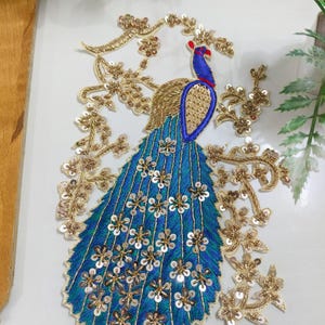 Pode incluir: Um aplique bordado de pavão com corpo azul e uma longa cauda em leque decorada com lantejoulas douradas e fio metálico. O pavão é rodeado por ramos bordados a ouro com detalhes de lantejoulas, criando um motivo floral detalhado.