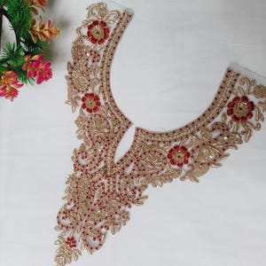 May include: A U-shaped neckline appliqué featuring intricate gold embroidery, metallic thread work, and small red floral accents. The design includes paisley-inspired swirls and beadwork, creating a decorative border for garment embellishment.