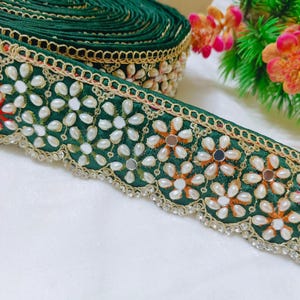 Può includere: Un rotolo di nastro in tessuto verde scuro con un motivo floreale ripetitivo creato con perle bianche finte e piccoli specchi circolari. I bordi sono rifiniti con un bordo in filo metallico dorato e una fila di cristalli trasparenti.