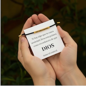 May include: A black cord bracelet with gold-colored accents and the text "DIOS TIENE EL CONTROL" on a gold bar. The bracelet is displayed on a white card with Spanish text that reads "DIOS tiene el control."