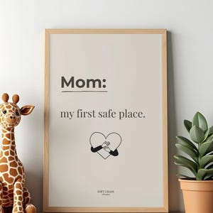 Puede incluir: Una impresión enmarcada con el texto "Mom: my first safe place." La obra de arte presenta un corazón con brazos que lo abrazan. Una jirafa de peluche y una planta en maceta están en un estante junto a la impresión enmarcada.