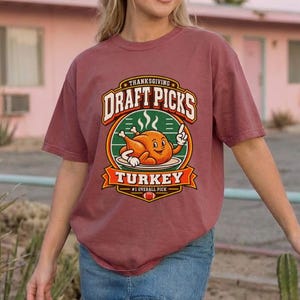 Puede incluir: Una camiseta de manga corta color rosa polvoriento con un gráfico de un pavo asado de dibujos animados en una bandeja con el texto "THANKSGIVING DRAFT PICKS TURKEY #1 OVERALL PICK" en un diseño de insignia circular con acentos naranja, blanco y verde.