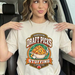 Puede incluir: Una camiseta de manga corta color crema con un estampado gráfico que incluye el texto "THANKSGIVING DRAFT PICKS STUFFING #1 OVERALL PICK". El diseño muestra un tazón de relleno de dibujos animados con cara, brazos y un icono de balón.