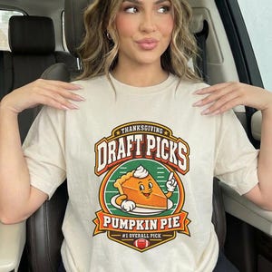 Puede incluir: Una camiseta de manga corta color crema con un estampado gráfico que incluye el texto "THANKSGIVING DRAFT PICKS" y "PUMPKIN PIE #1 OVERALL PICK". El diseño muestra una porción de tarta de calabaza con cara, brazos y un balón de fútbol.