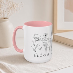 May include: White ceramic mug with a pink interior and handle. The mug features a black line drawing of various flowers and the word "BLOOM" in black letters. The mug is sitting on a white cloth.