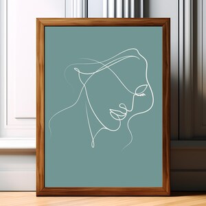Op de afbeelding: Een ingelijste muurkunstprint met een doorlopende witte lijntekening van een menselijk gezicht in profiel tegen een effen, gedempte blauwgroene achtergrond. Het kunstwerk zit in een rechthoekige houten lijst met een bruine afwerking.
