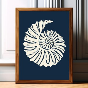 May include: A framed wall art print featuring a cream-coloured, stylised illustration of a spiral seashell centred on a solid dark navy blue background. The artwork is enclosed in a simple, medium-toned wooden frame with a clean, rectangular profile.
