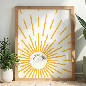 May include: A framed print featuring a yellow sunburst design on a white background. The words "positive rays" are in a circle at the center. The wooden frame is light brown. Two potted plants flank the artwork.