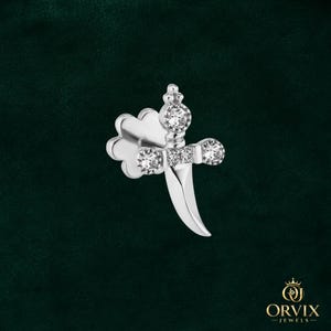 May include: A silver-toned earring shaped like a small dagger with a curved blade. The hilt features three clear, round-cut stones and a small clover-shaped backing. The design is metallic and polished, with a sharp, pointed tip at the bottom.