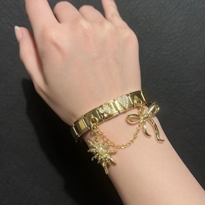 Puede incluir: Pulsera dorada con varios dijes, incluyendo un lazo, un sol, un corazón y una flor. La pulsera es rectangular y tiene un cierre. Los dijes están unidos con pequeñas cadenas. La pulsera se lleva en la muñeca.