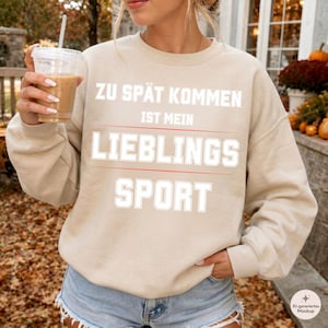 Könnte beinhalten: Ein beigefarbener Rundhals-Pullover mit dem weißen Schriftzug ZU SPÄT KOMMEN IST MEIN LIEBLINGS SPORT in fetter Blockschrift auf der Brust, unterstrichen und getrennt durch dünne, horizontale rote Linien.