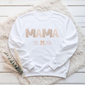 Könnte beinhalten: Weißes Sweatshirt mit dem Wort "MAMA" in Buchstaben mit tanfarbenen Punkten und Vichy-Karo-Muster. Unter dem Wort "MAMA" steht der Text "EST. 2026" mit einer Schleifengrafik. Das Sweatshirt wird auf einer weißen, flauschigen Oberfläche präsentiert.