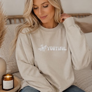 Könnte beinhalten: Ein beigefarbenes Sweatshirt mit Rundhalsausschnitt und weißer Stickerei auf der Brust. Das Design zeigt eine Biene mit einer kleinen Krone links neben dem Wort YOUTIFUL, das in einer abgerundeten, blasenartigen Schriftart geschrieben ist.