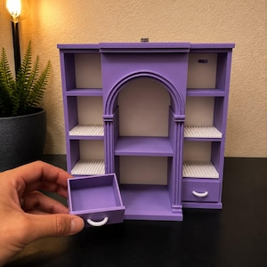 May include: A purple miniature shelving unit with a central arched alcove, multiple open shelves, and two small drawers with white handles. One drawer is pulled out, revealing its hollow interior. The structure features a smooth, matte finish.