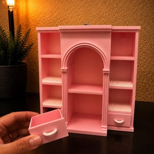 May include: A pink miniature shelving unit with an arched central section, two side columns featuring open shelves, and two small drawers with white handles. One drawer is detached and held in front of the unit, revealing white ribbed shelf liners.