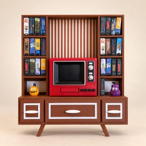 May include: A brown miniature entertainment center with a red television set, shelves filled with small VHS tapes, and two small pumpkin figurines. The unit features a central drawer, two side cabinets with white trim, and a striped back panel.